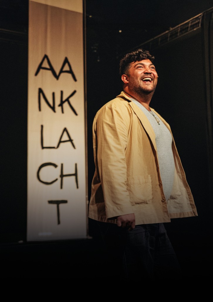 Een man staat op een podium, lachend en kijkend naar boven. Hij draagt een beige jas en een grijs shirt. Achter hem hangt een verticale banner met de tekst